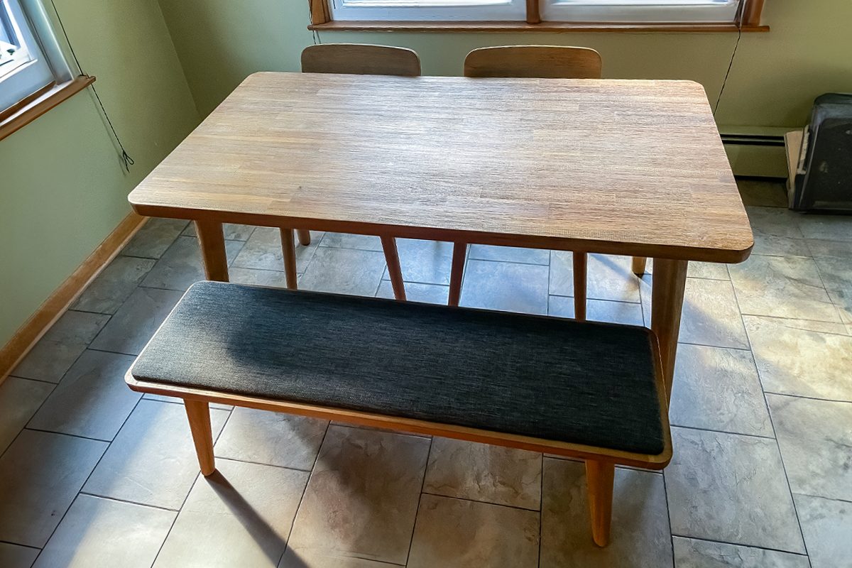 A wooden dining table with a bench on one side and two chairs on the other, set on a tiled floor in a sunlit room with windows in the background.