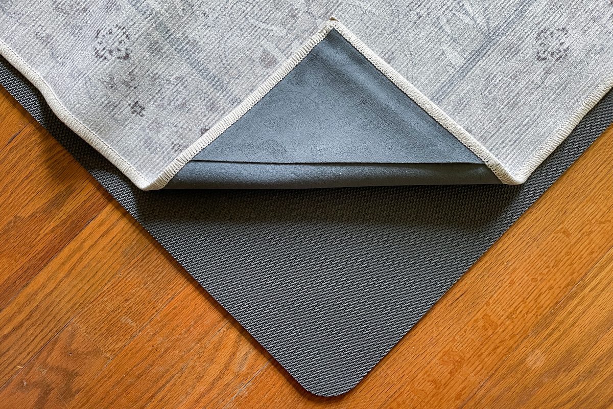 A corner of a patterned rug is lifted to reveal a gray non-slip rug pad underneath, both placed on a wooden floor.