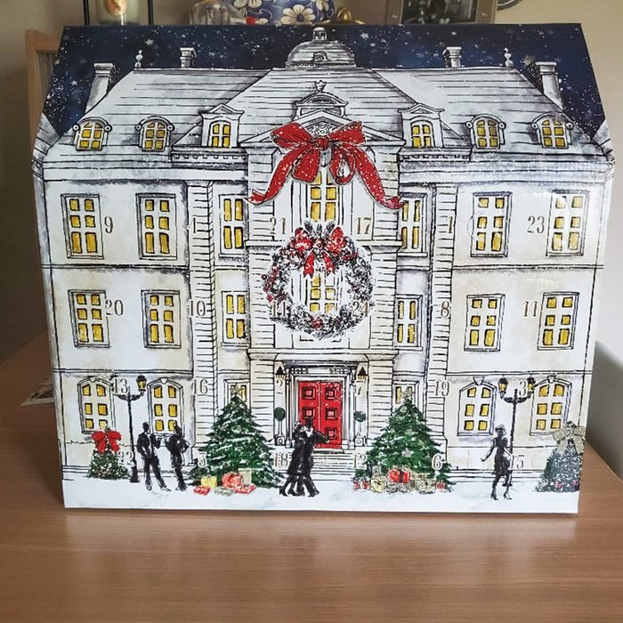 A decorative advent calendar shaped like a grand, snow-covered house with a red door, wreath, and large red bow. People in winter clothing stand by Christmas trees and lamp posts in front.