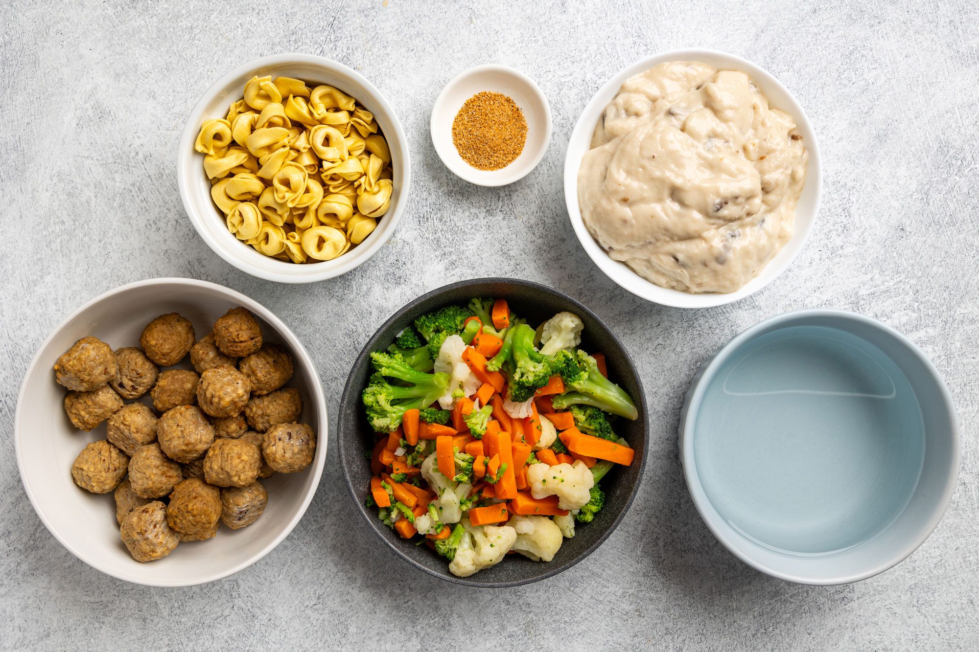 Overhead shot; bowls of tortellini; meatballs; steamed vegetables; creamy sauce; breadcrumbs; empty bowl on light gray background
