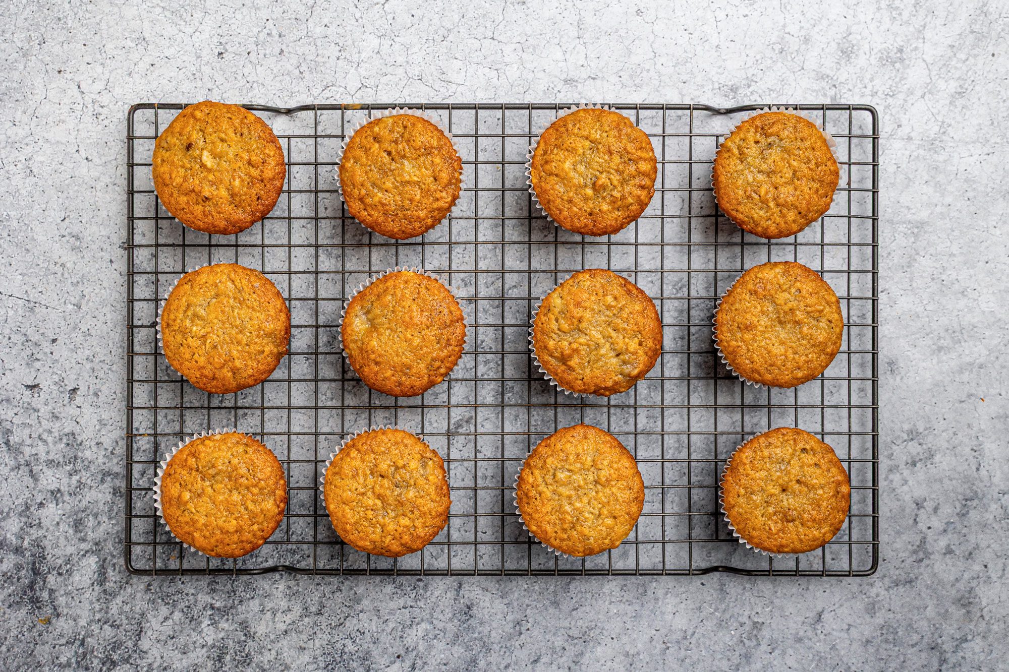 Overhead shot of twelve golden-brown muffins arranged in rows on a wire cooling rack, set on a gray textured surface