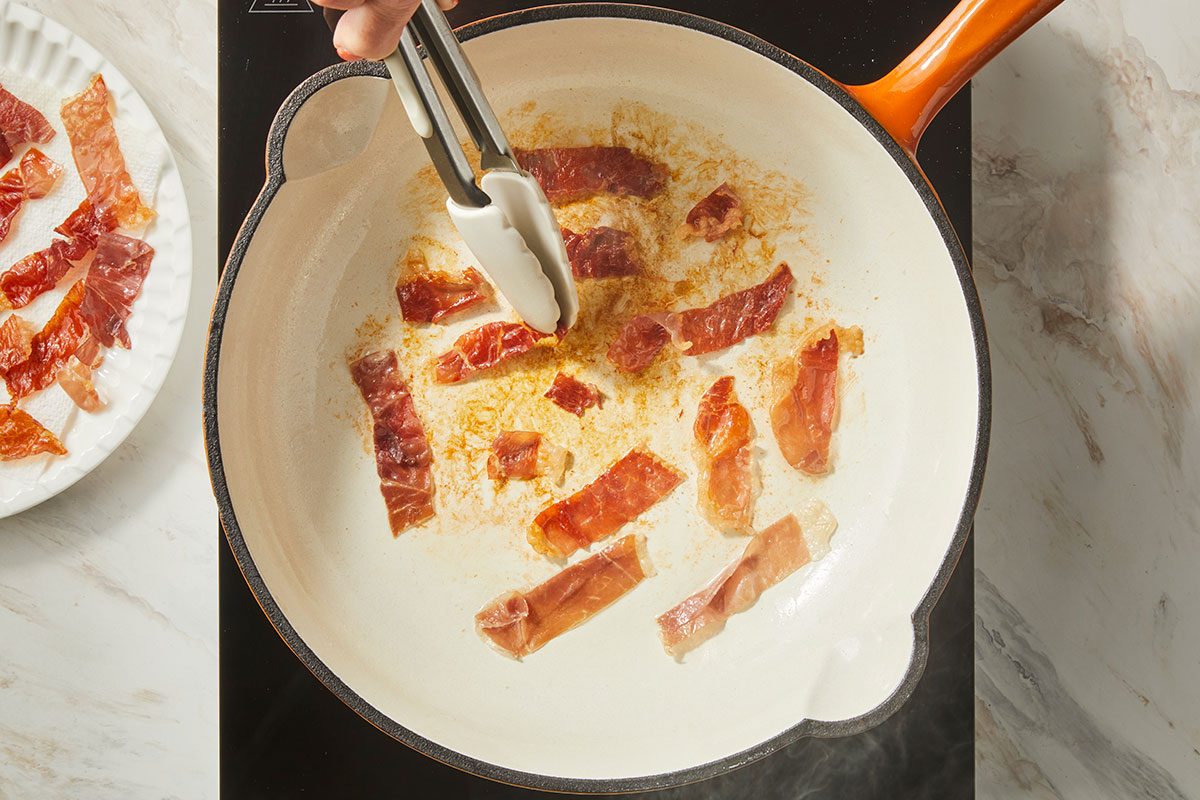 overhead shot of a hand uses tongs to cook strips of prosciutto in a white skillet on a stovetop, with more prosciutto pieces on a plate nearby