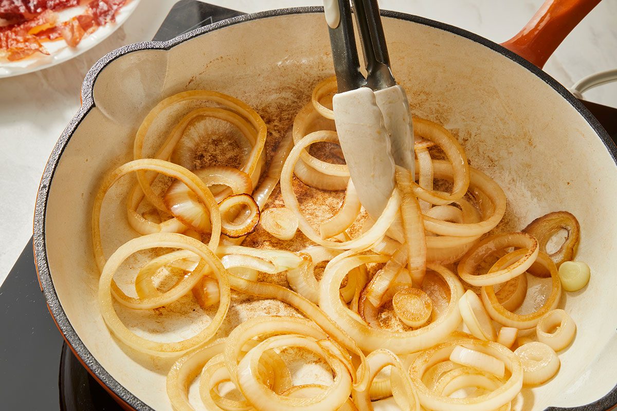 3/4th shot of a pair of tongs stirs sliced onions in a white cast iron skillet on a stovetop; cooked bacon is visible on a plate in the background
