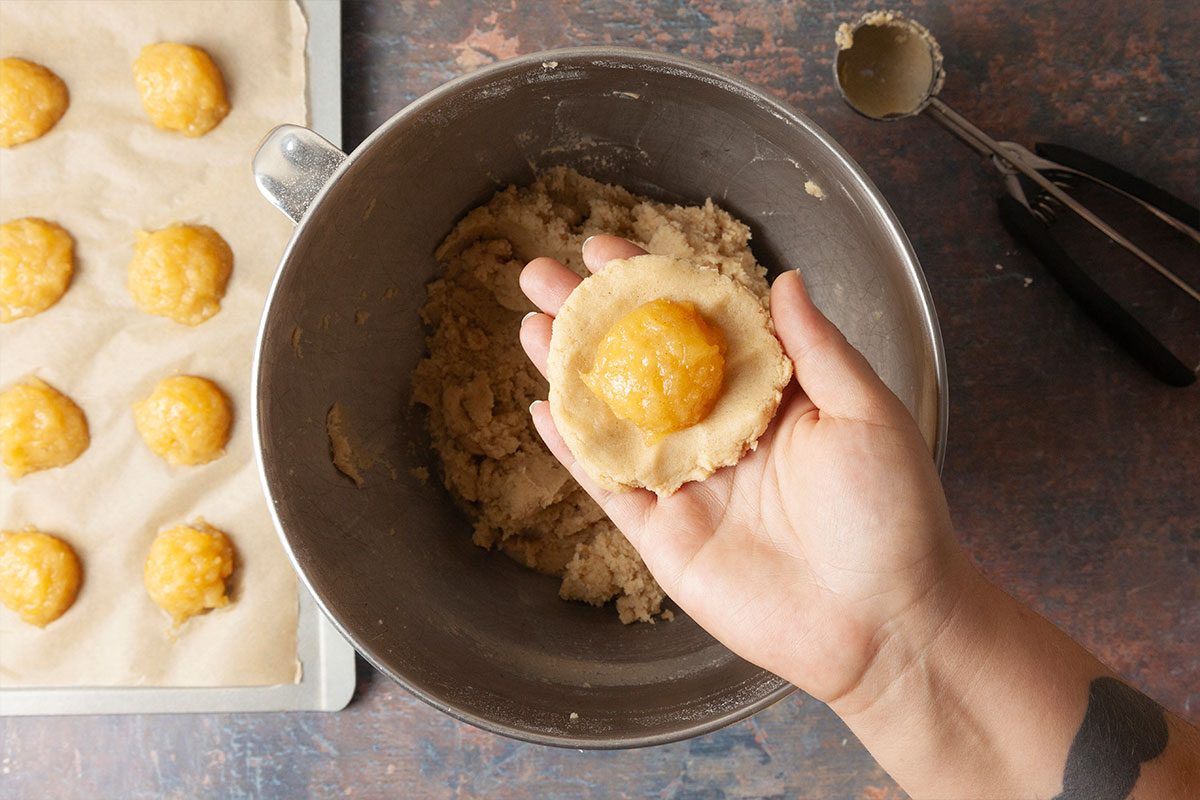 Overhead shot of a hand holding a piece of cookie dough with yellow jam in the center over a mixing bowl, with a baking tray of similar jam-filled cookies nearby