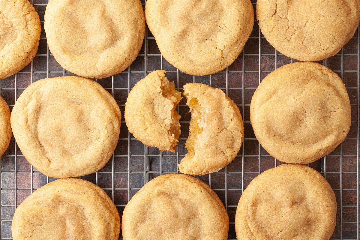 Overhead shot of several golden-brown Apple Pie-Stuffed Cookies cooling on a wire rack, with one broken in the center to reveal its soft, gooey interior