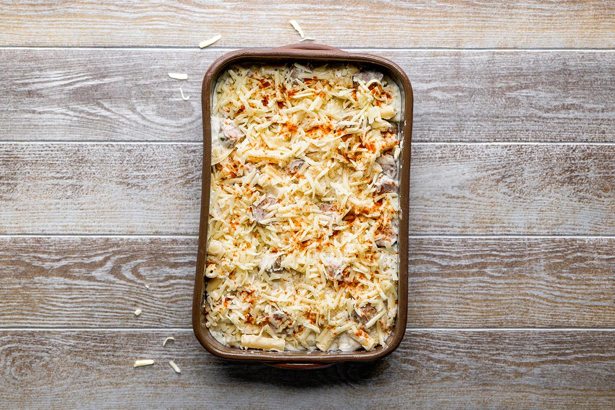 A rectangular baking dish filled with an uncooked casserole topped with shredded cheese and a sprinkle of seasoning, sitting on a wooden surface.