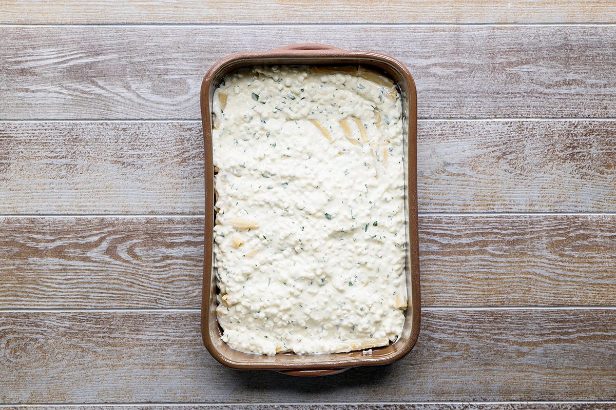 A rectangular baking dish filled with an unbaked lasagna, topped with a thick layer of creamy white cheese mixture, sits on a wooden surface.