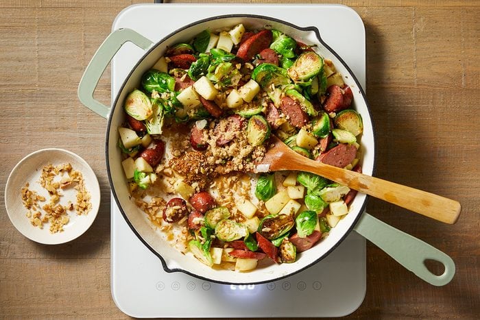 A skillet on a stove filled with chopped Brussels sprouts, sausage, apples, and walnuts being stirred with a wooden spoon; a small bowl of walnuts sits nearby on the wooden countertop.