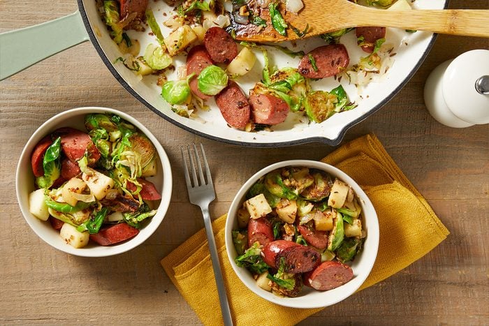 Two bowls of sautéed sausage, potatoes, and Brussels sprouts, with a pan of the same dish nearby. A fork rests on a yellow napkin beside the bowls, all set on a wooden table.