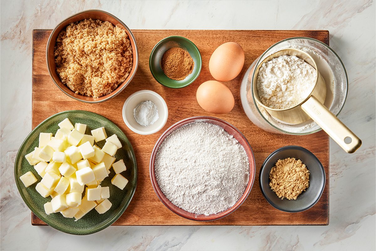 Overhead shot of a wooden board with bowls of brown sugar, cubed butter, flour, baking powder, cinnamon, two eggs, grated ginger, and a sifter with flour on a marble surface, ready for baking;