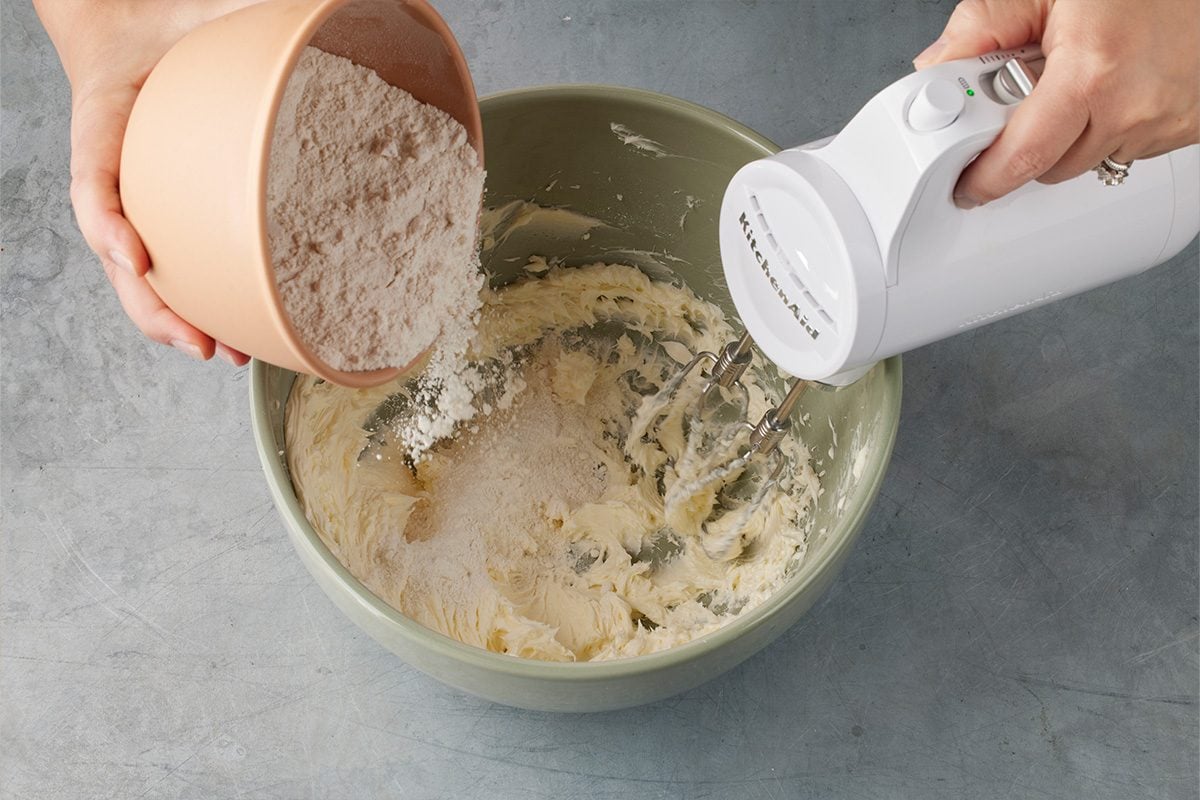 Overhead shot of a person pouring flour from a bowl into a green mixing bowl containing creamed ingredients, while using a white electric hand mixer on a gray countertop;