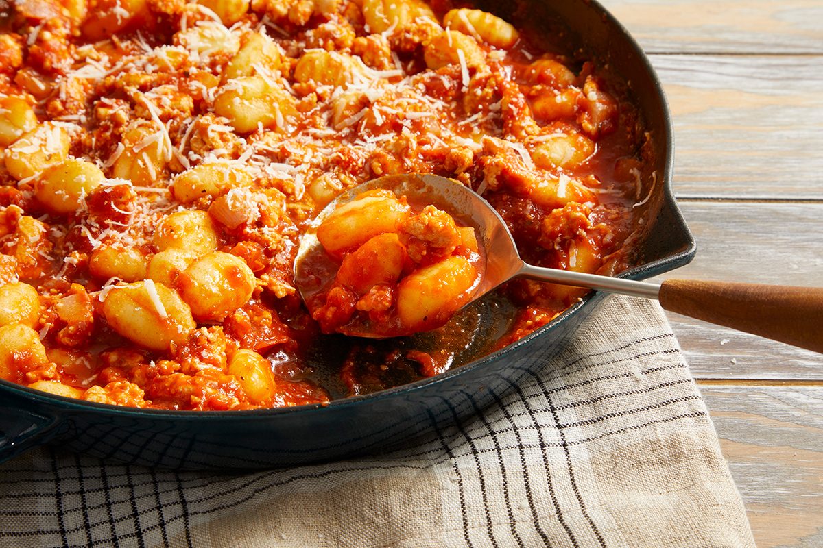A skillet filled with gnocchi in a rich tomato sauce with crumbled sausage, topped with grated Parmesan cheese. A spoon is scooping some of the dish, resting on a striped cloth.