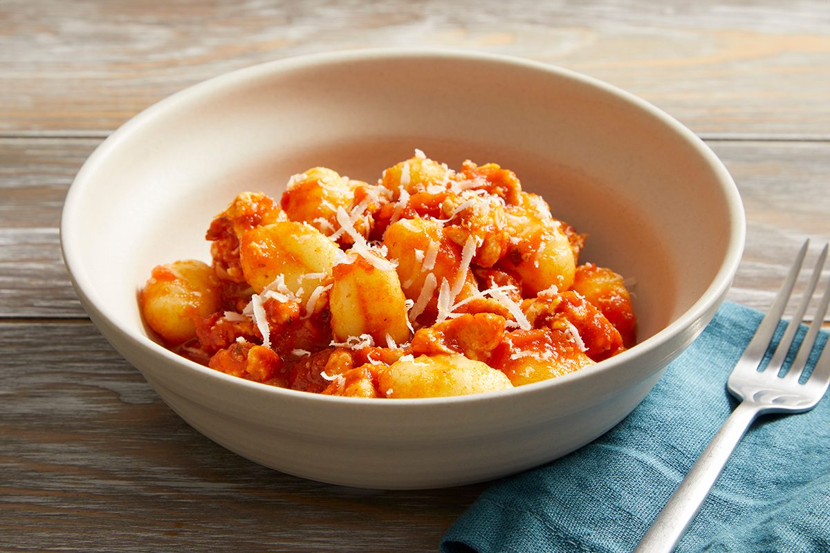 A white bowl filled with gnocchi in tomato sauce, topped with grated cheese, sits on a wooden table next to a fork and a blue napkin.
