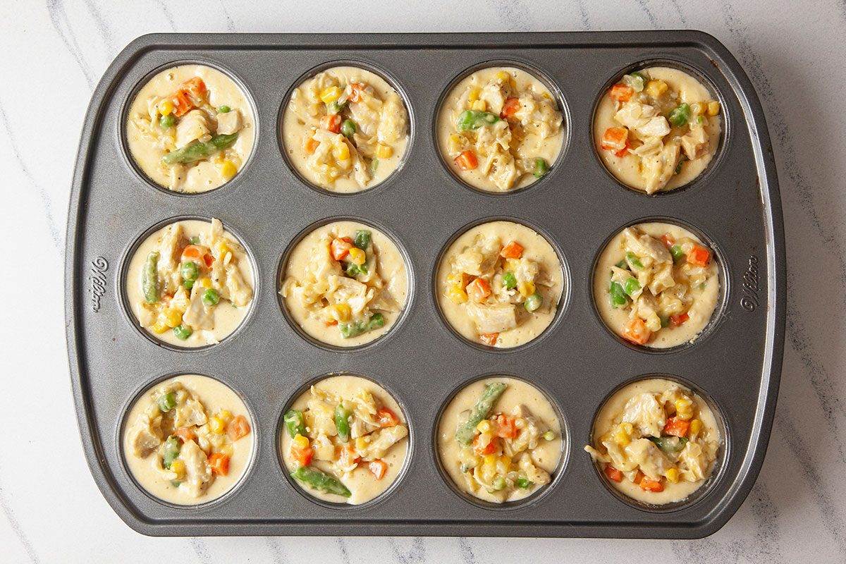 Overhead shot of a muffin tin with 12 cups filled with an uncooked mixture of eggs, vegetables, and chicken, ready to be baked; ingredients include corn, green beans, carrots, and peas;