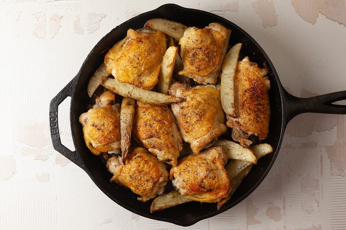 Overhead shot of a cast-iron skillet filled with baked, golden-brown chicken thighs and potato wedges, neatly arranged on a light background
