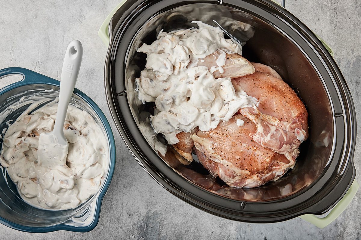 Raw chicken breasts in a slow cooker are topped with a creamy mushroom mixture. Next to the slow cooker, a blue bowl holds more of the creamy mixture with a white spatula resting inside.