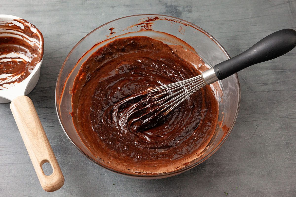 3/4th shot of a glass bowl is being used to mix chocolate cake batter with a whisk on a dark countertop and a white spatula with a wooden handle covered in batter rests nearby