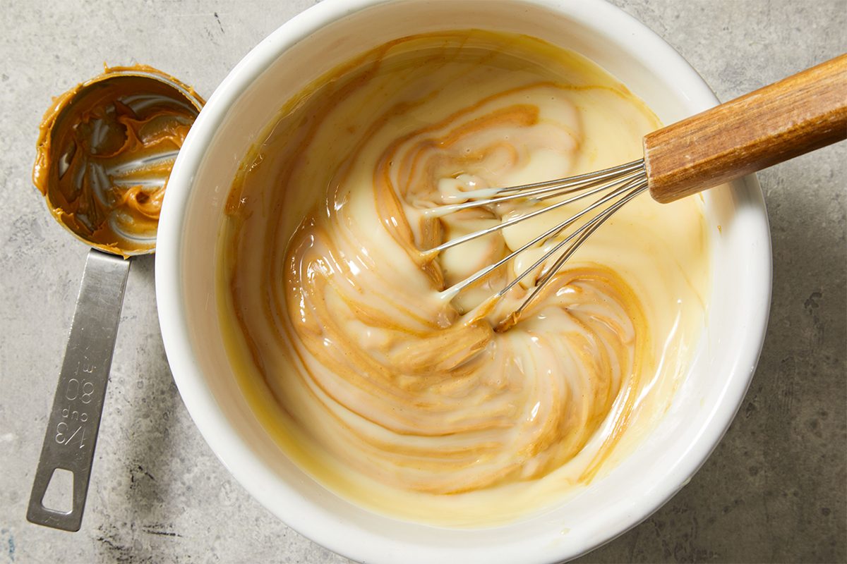 A metal whisk stirs a creamy mixture of caramel and sweetened condensed milk in a white bowl, creating a marbled swirl effect. A measuring cup with caramel residue sits nearby on a textured surface.