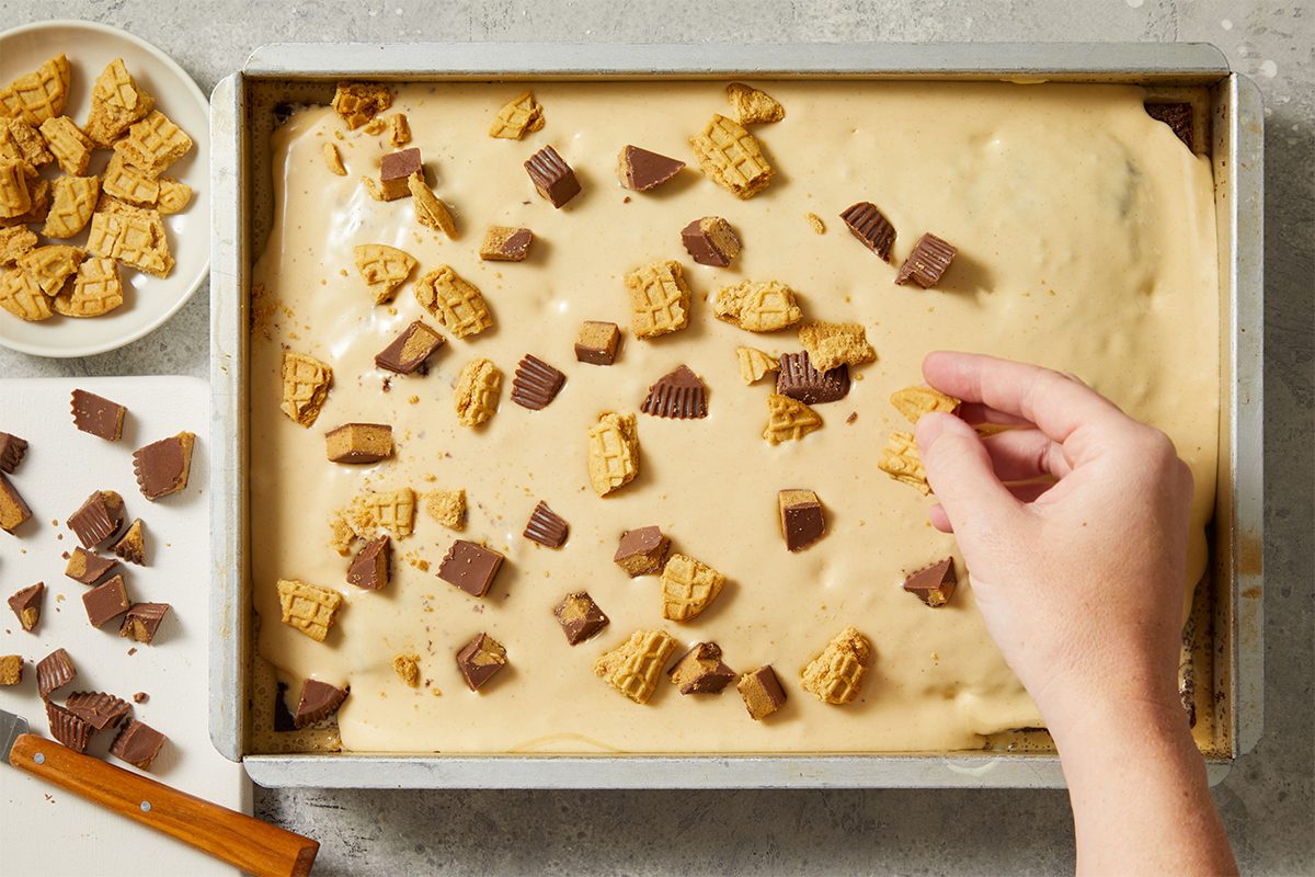 A hand sprinkles chopped peanut butter cups and broken cookies onto a pan of light brown batter, with extra toppings on a small plate and cutting board nearby. The scene is set on a light grey surface.