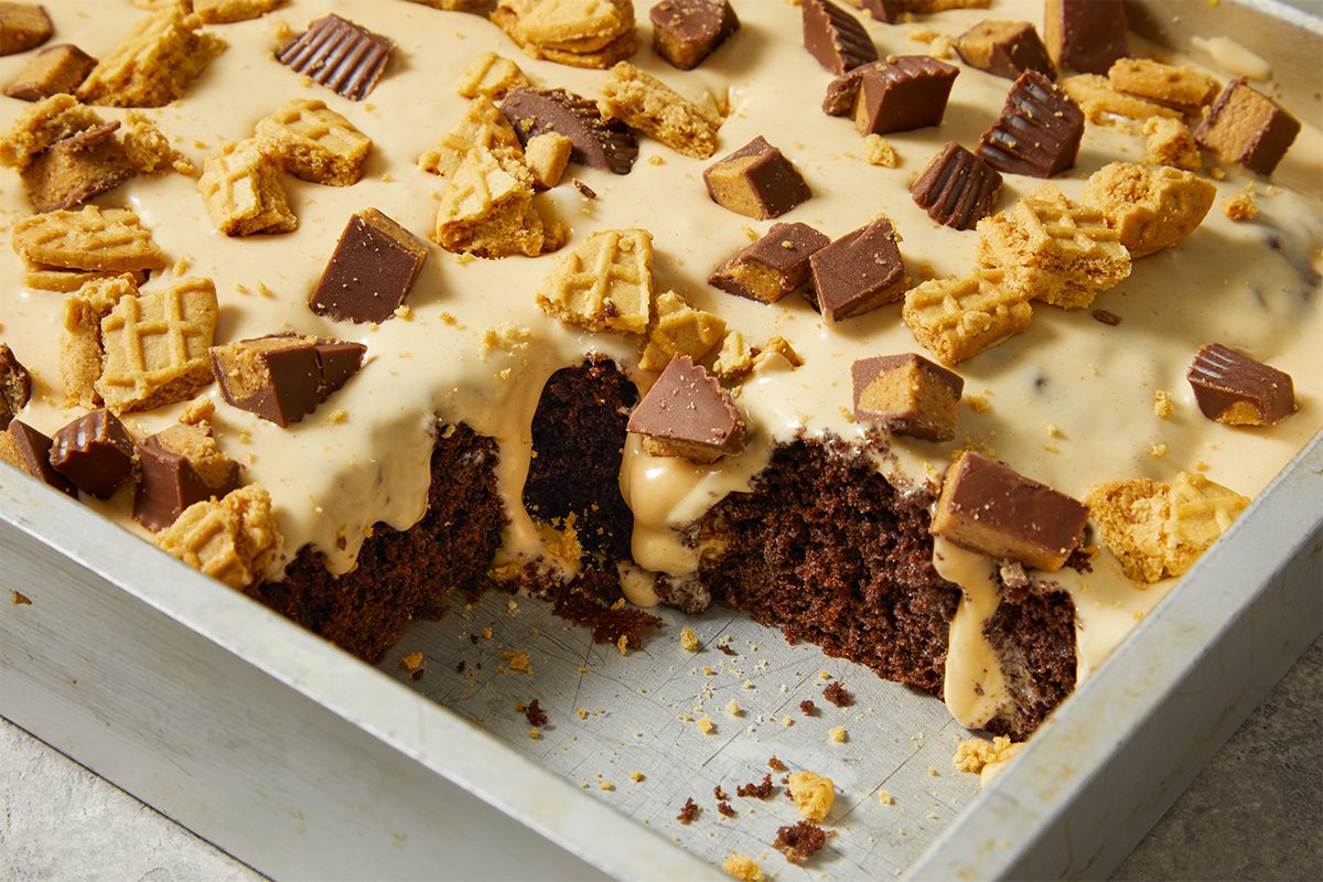 A chocolate sheet cake in a metal pan is topped with tan frosting, chopped peanut butter cups, and crumbled cookies. A large square piece has been cut and removed, showing the moist cake inside.