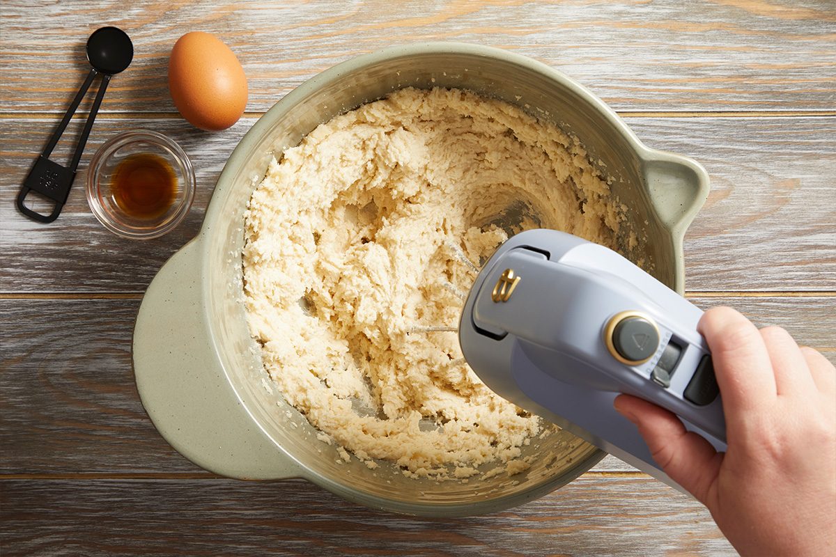 Overhead shot of an electric hand mixer blending cookie dough in a light green mixing bowl with eggs and vanilla nearby on a wooden table.