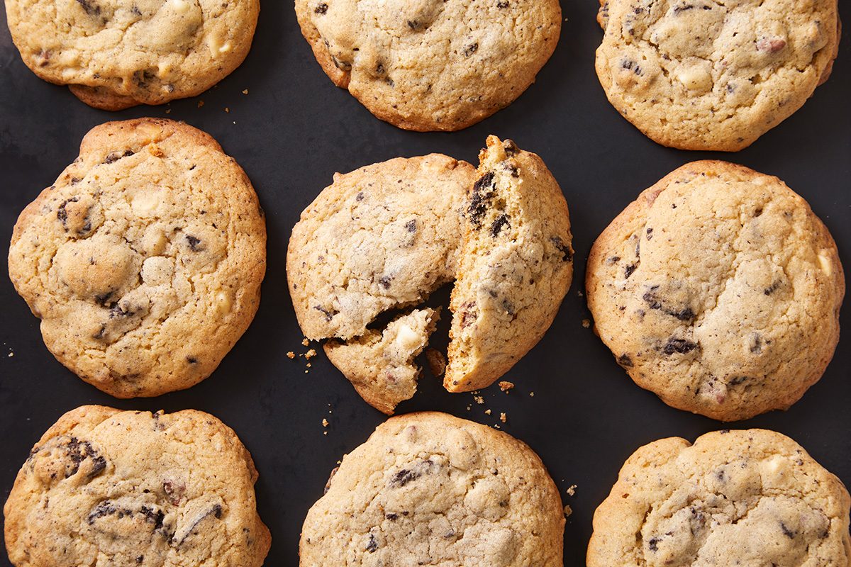 Nine chocolate chip cookies are arranged in a grid on a dark surface. The center cookie is broken in half, showing its soft interior and some crumbs. The cookies are golden brown and look freshly baked.