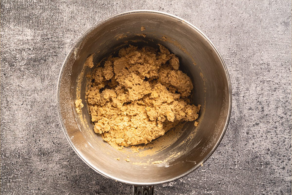 A metal mixing bowl containing a rough, light brown cookie dough mixture sits on a textured gray surface.