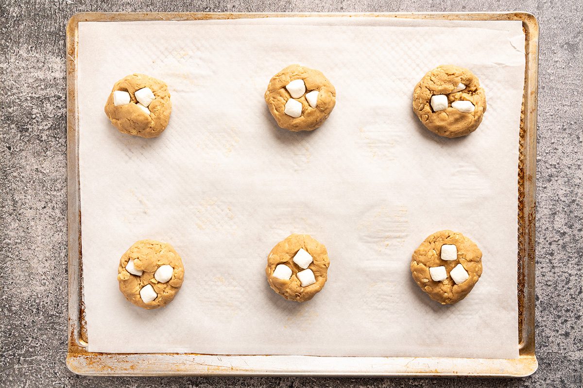 Six unbaked cookies with white marshmallow pieces on top are evenly spaced on a parchment-lined baking sheet, ready to be baked. The background is a textured gray surface.