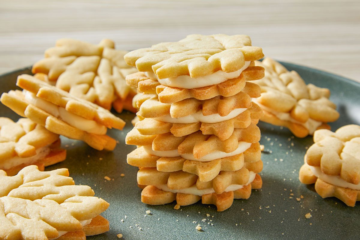 Horizontal Shot of Copycat Trader Joe's Maple Leaf Cookies are stacked on a dark plate and creamy filling peeks out more cookies rest on a light wood surface