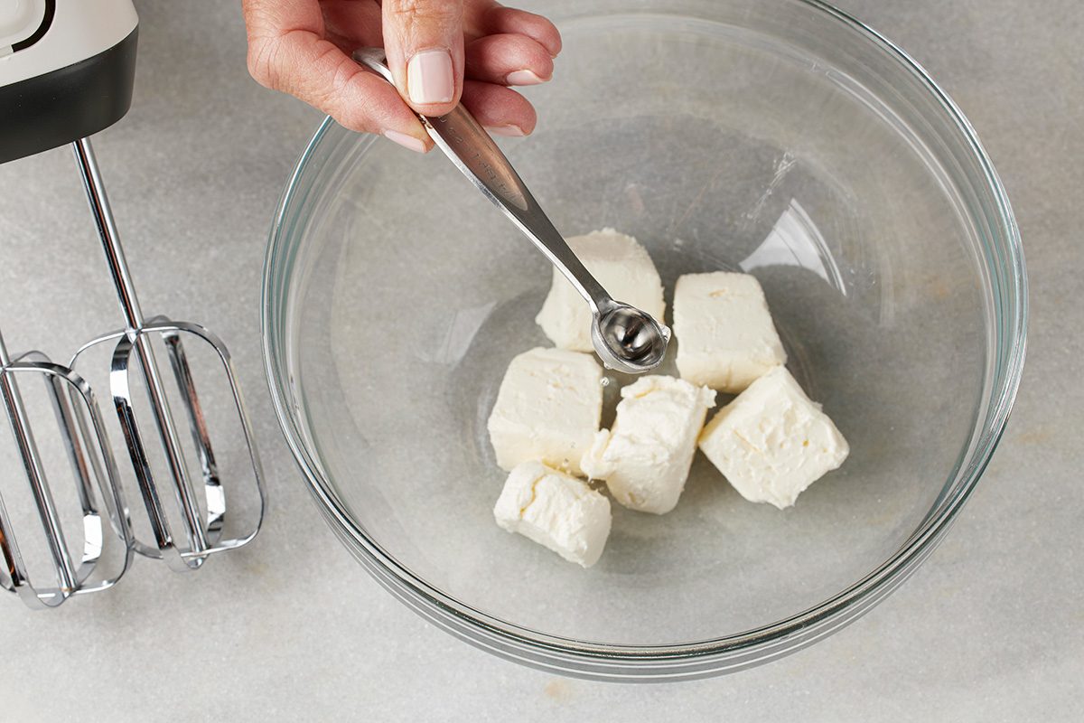 Overhead shot of a hand holding a measuring spoon over a glass bowl with cubes of cream cheese