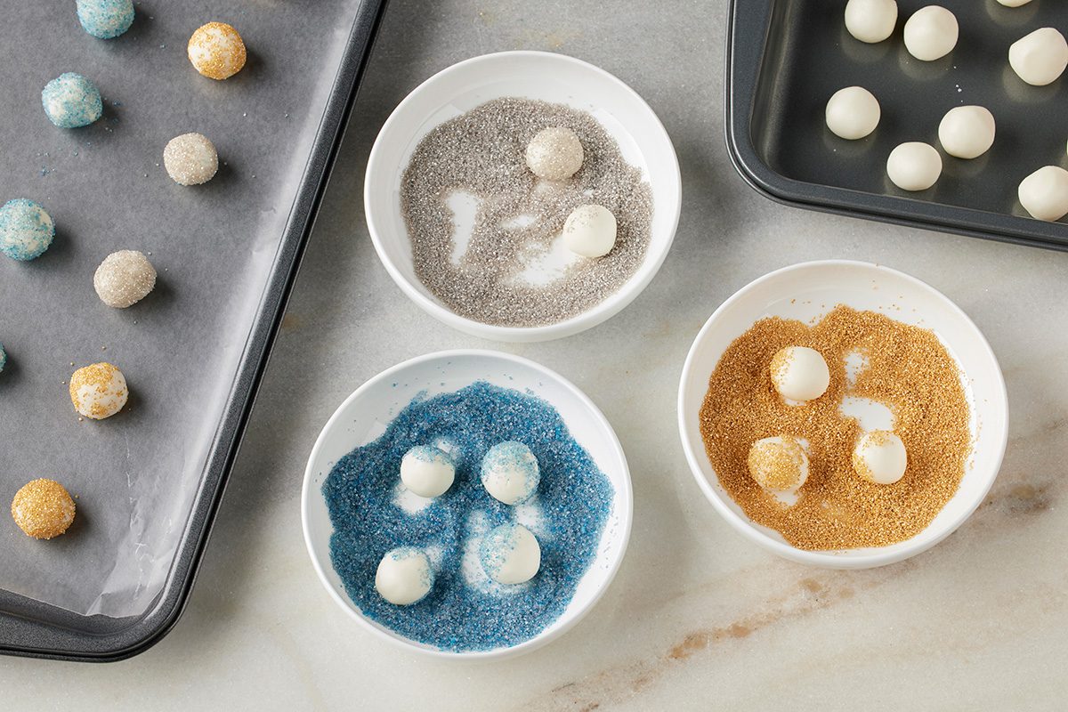 Overhead shot of three bowls filled with blue, silver, and gold sugar used to coat small white dough balls