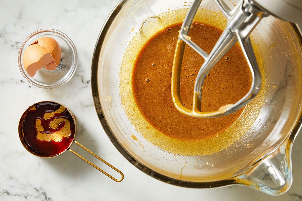 Overhead shot of a glass mixing bowl holds brown batter with a mixer beside it and a small bowl has a broken eggshell and there is a cup of molasses nearby all on a white marble surface