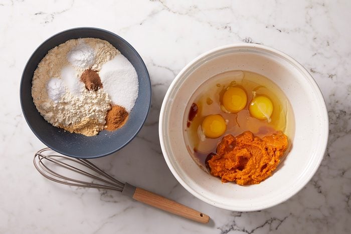 Two mixing bowls on a marble surface: one with flour, spices, sugar, and baking soda; the other with eggs, pumpkin puree, and vanilla. A whisk lies beside the bowls.