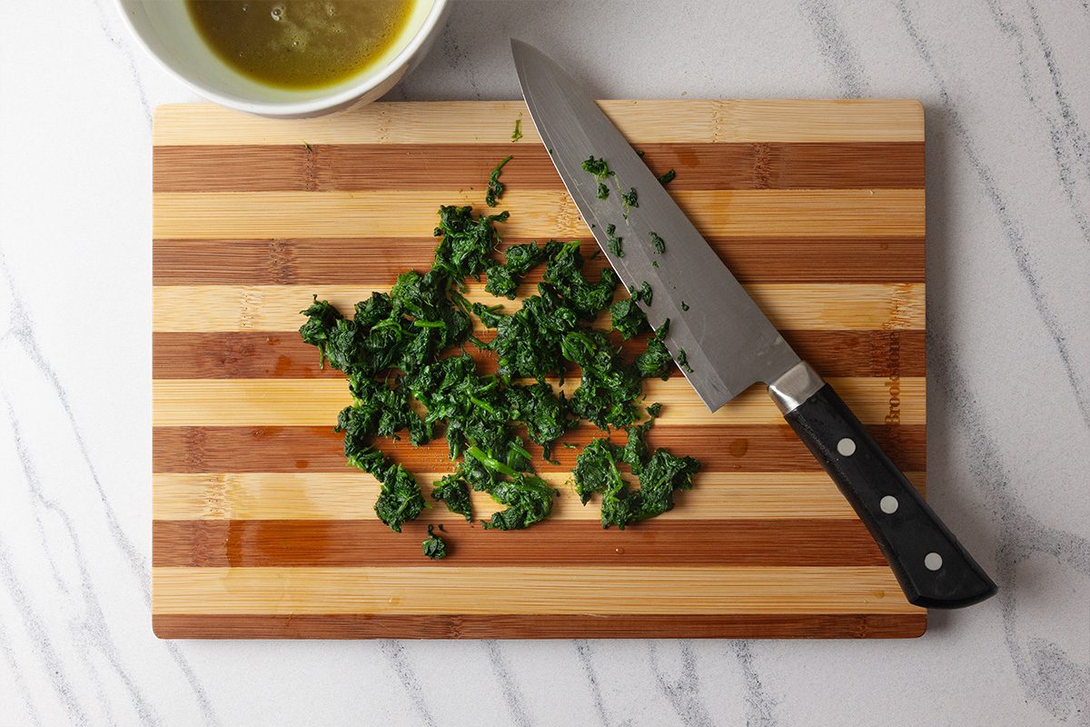 Overhead shot of a chef’s knife and chopped parsley on a striped wooden cutting board, with a bowl of green liquid nearby on a light marble countertop