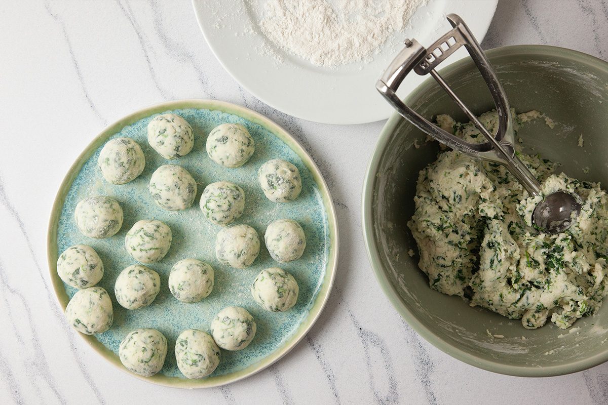 Overhead shot of a plate of uncooked spinach and ricotta dumplings beside a bowl of dough and a scoop on a white countertop, with a plate of flour in the background