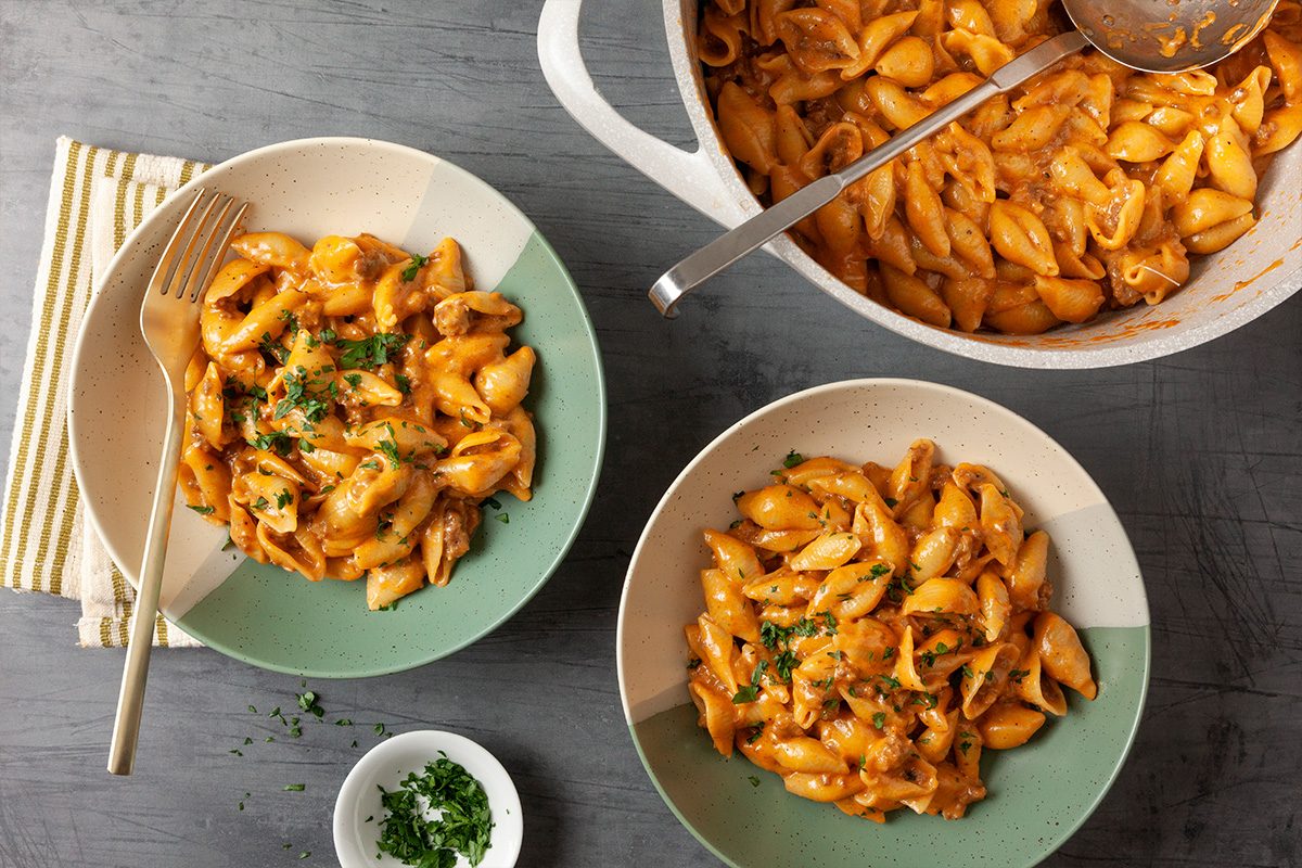 Overhead shot of Homemade Hamburger Helper in two bowls with herbs sits beside a pot of more pasta Fork and napkin are on the gray table plus extra herbs