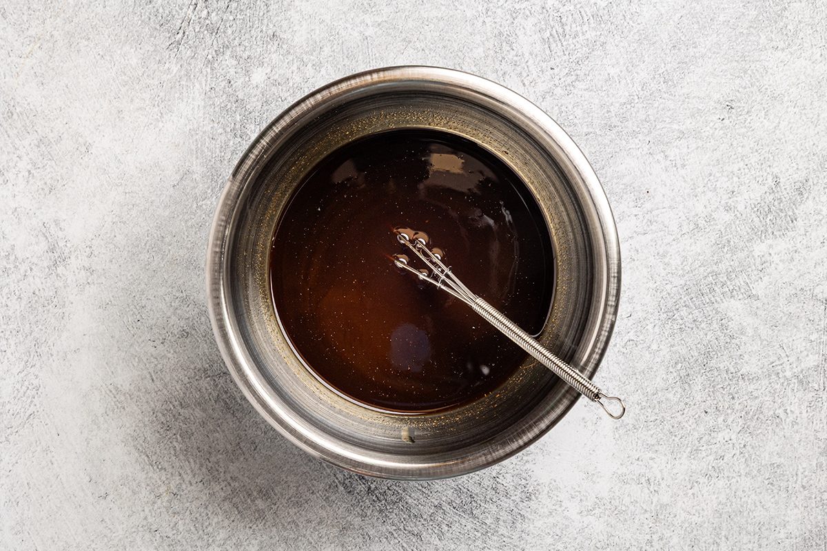 A metal bowl filled with a dark liquid sits on a light gray textured surface. A small metal whisk rests inside the bowl.