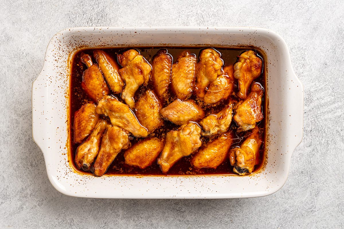A white rectangular baking dish filled with glazed baked chicken wings in a rich, reddish-brown sauce, set on a light textured surface.
