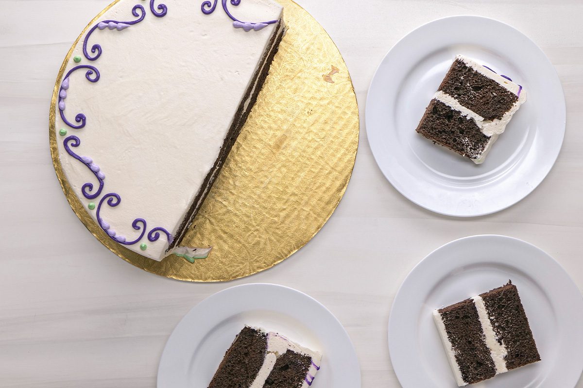 A round chocolate layer cake with white frosting and purple decorations sits on a gold cake board. Three slices are served on white plates beside it.