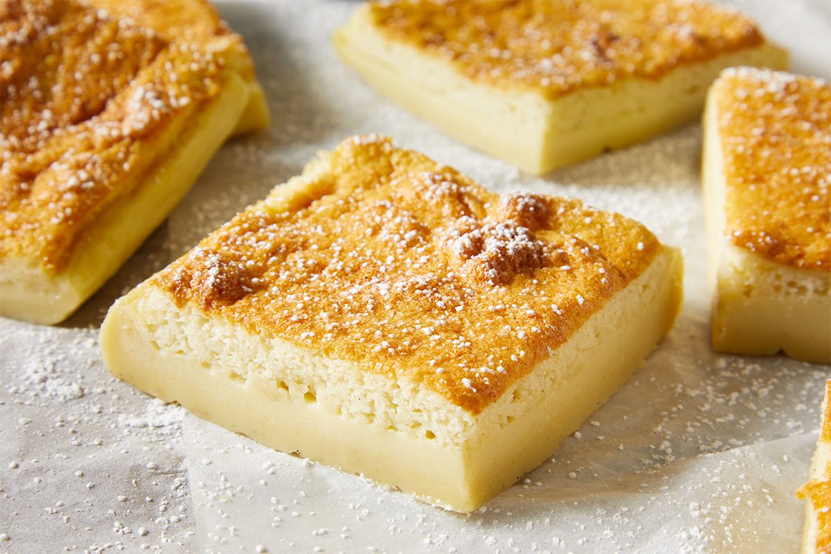 Squares of golden, slightly browned custard cake sit on parchment paper, dusted with powdered sugar. The cakes have a dense, creamy bottom layer and a light, airy top layer.