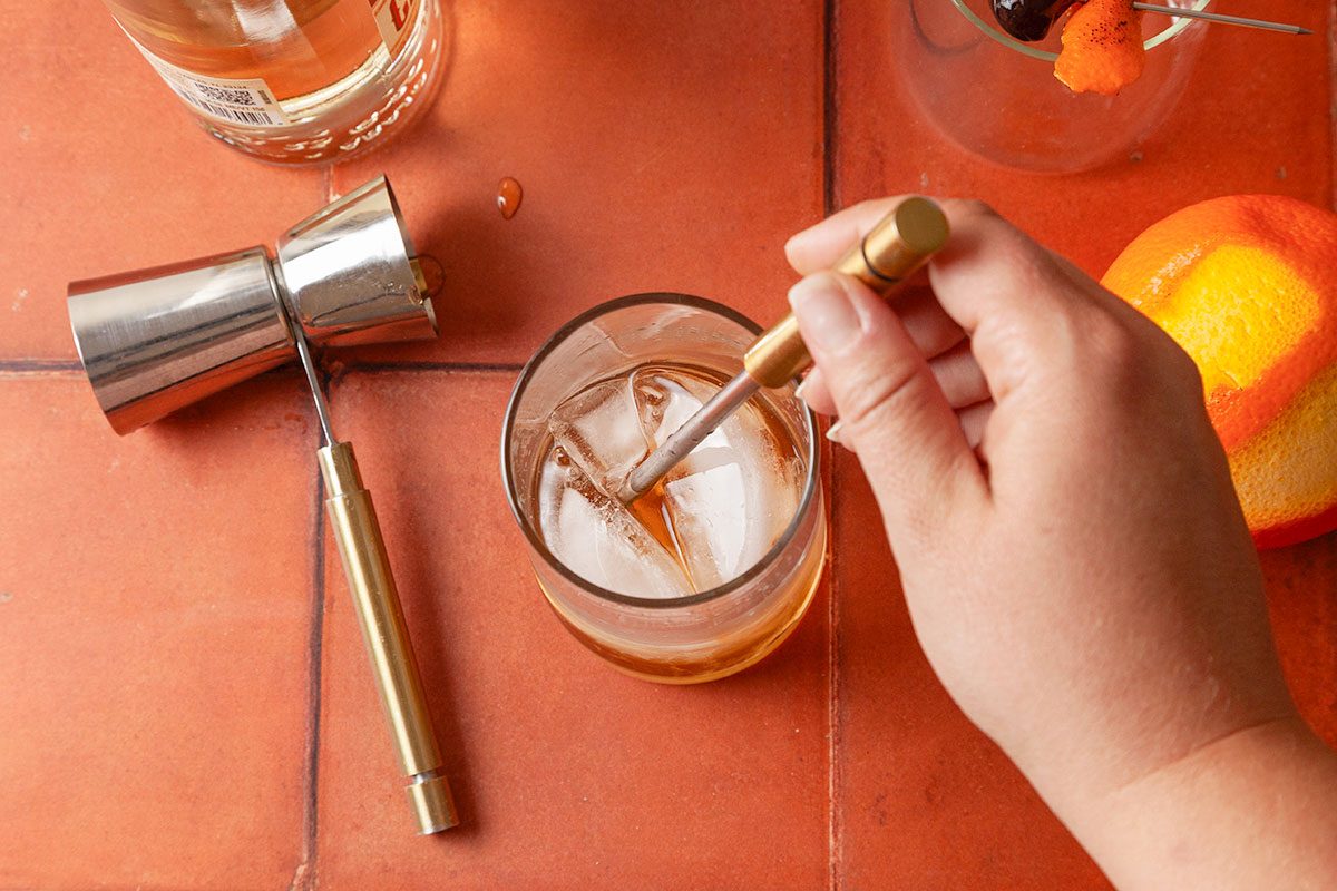 Overhead shot of a hand stirring a cocktail in a glass filled with ice using a metal stirrer