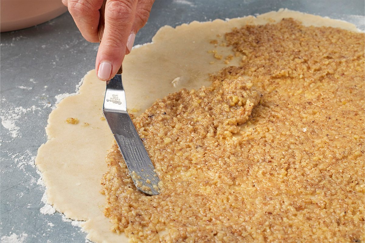 Close-up of a hand using a metal spatula to spread a nutty filling over rolled-out dough on a gray surface, preparing for baking;