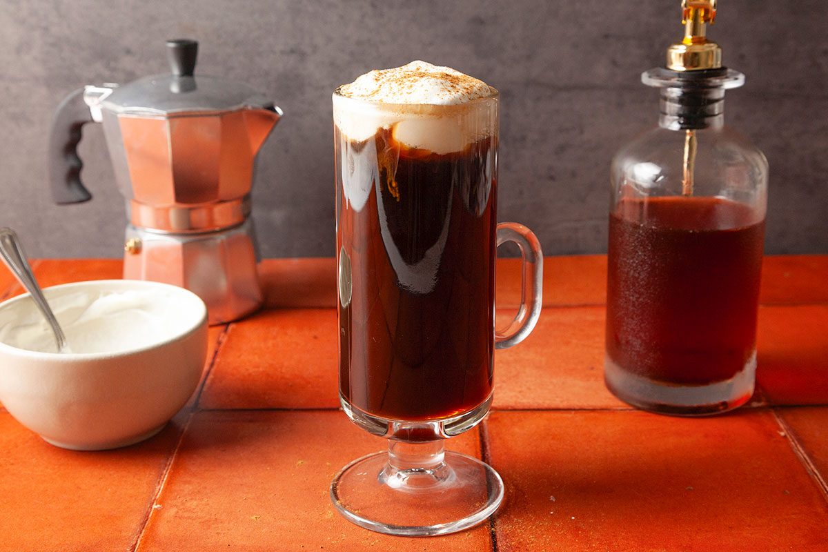 Table view shot of a glass mug of coffee topped with frothy cream on a reddish tile surface, with a bowl of cream, a moka pot, and a bottle of syrup in the background;