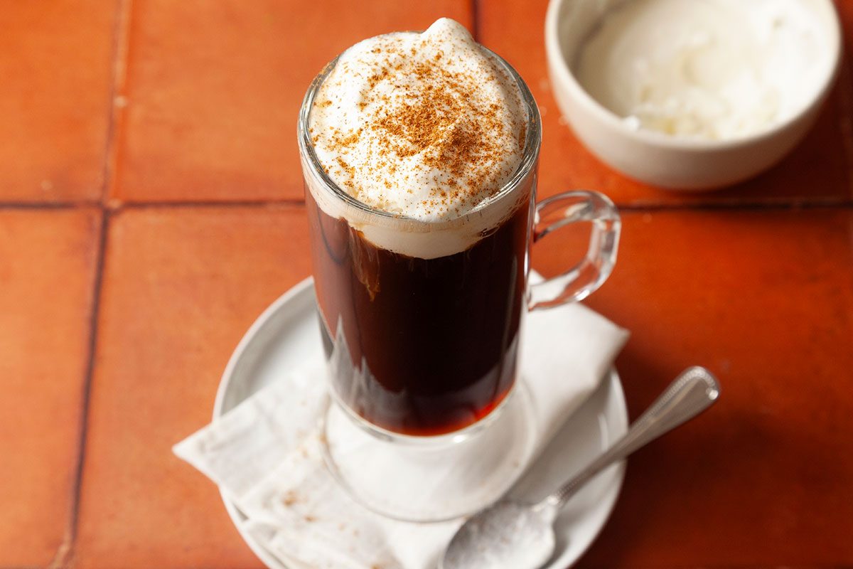 Oaxacan Coffee in a glass mug with whipped cream and cinnamon sits on a plate with a spoon, bowl of cream, and terracotta tiles;