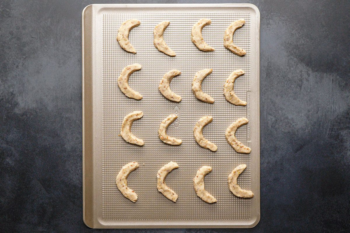Overhead Shot; A baking sheet sits on a dark countertop holding 18 unbaked crescent-shaped cookies arranged neatly in rows and ready to bake