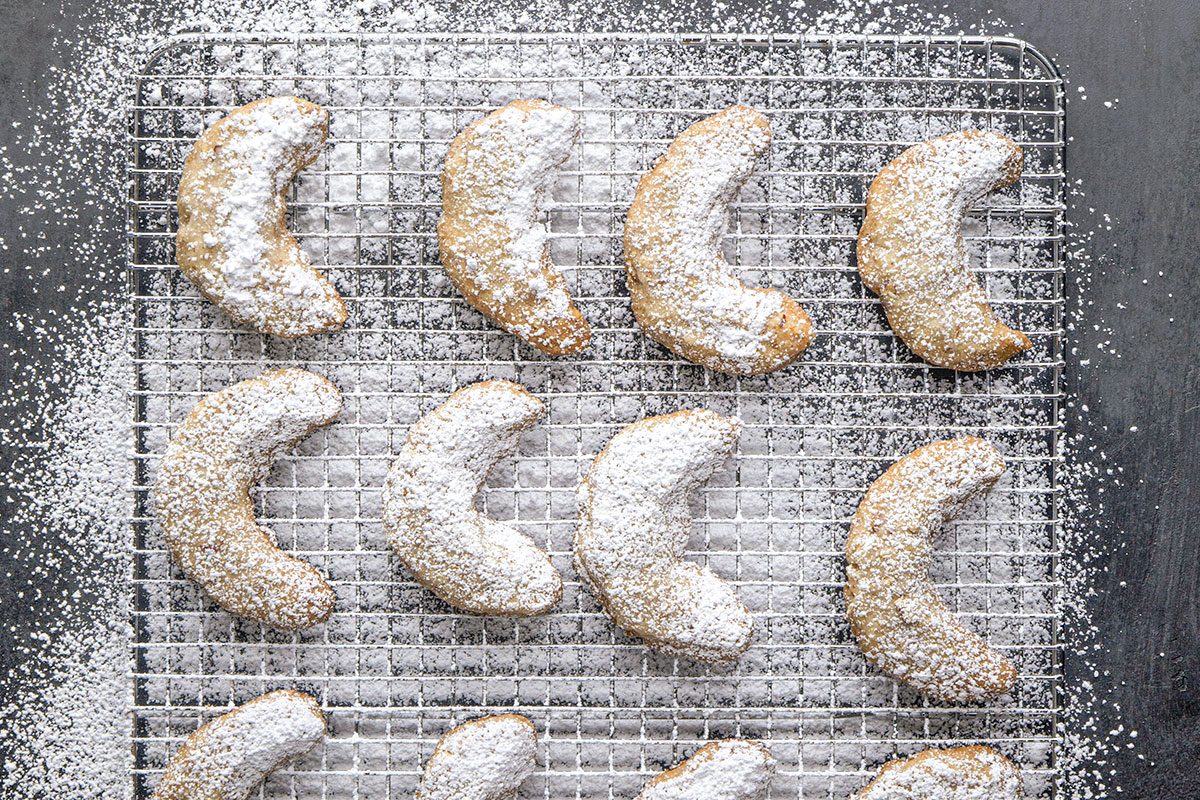 Overhead Shot; Powdered sugar-dusted crescent-shaped cookies sit lined up in rows on a cooling rack and extra sugar sprinkles all around them