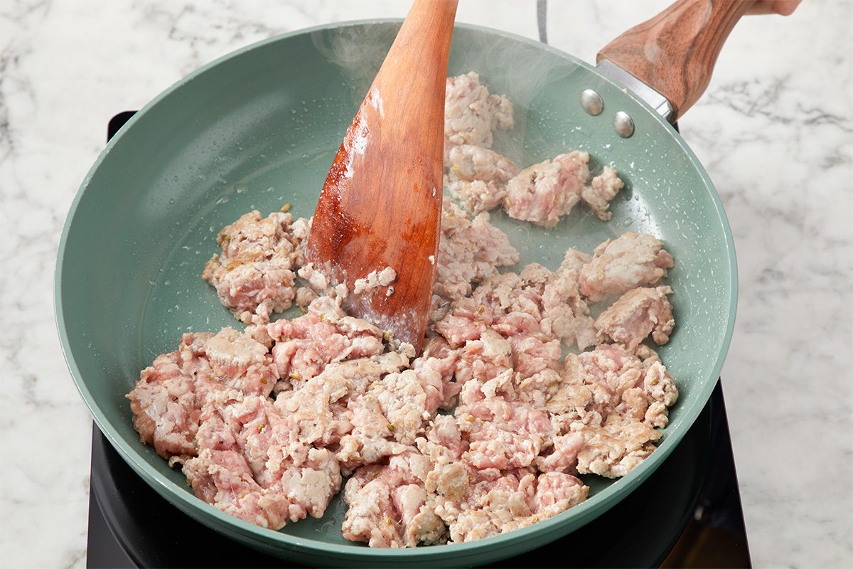 Ground meat is being cooked and stirred in a green nonstick frying pan with a wooden spatula on a stovetop. The meat is partially browned, and the pan sits on a marble countertop.