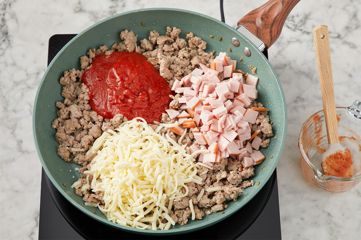 A skillet on a stovetop contains cooked ground meat, shredded cheese, diced ham, and tomato sauce, arranged in separate sections, ready to be mixed together. A glass measuring cup and wooden spoon are nearby.