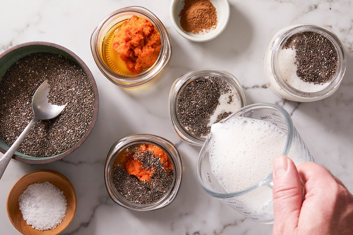 A hand pours milk into a jar with chia seeds on a marble countertop. Other jars hold chia seeds, pumpkin puree, and spices, with a bowl of salt and a spoon nearby.