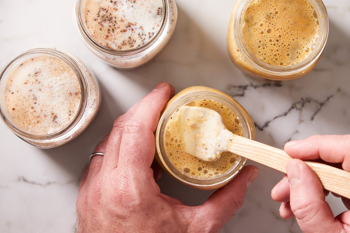 A hand holds a wooden spoon, stirring a frothy liquid in a small glass jar, with three other jars of foamy liquids nearby on a marble surface.